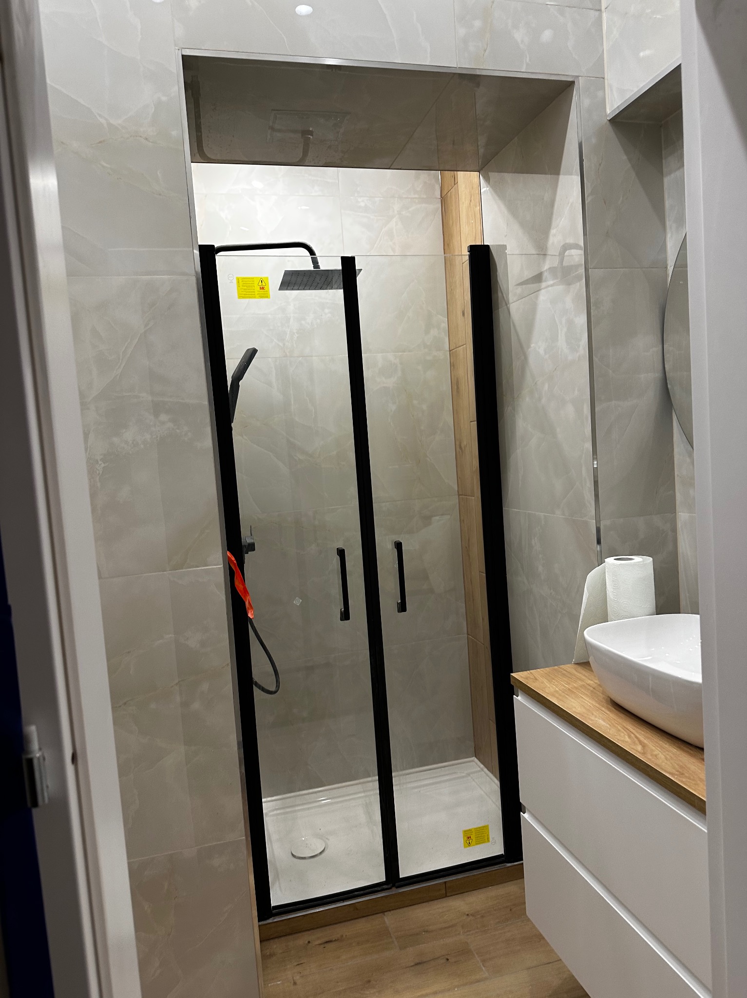 Nowoczesna kabina prysznicowa z czarnymi ramami i deszczownicą w łazience wykończonej płytkami imitującymi marmur, widoczna umywalka nablatowa i drewniany blat szafki