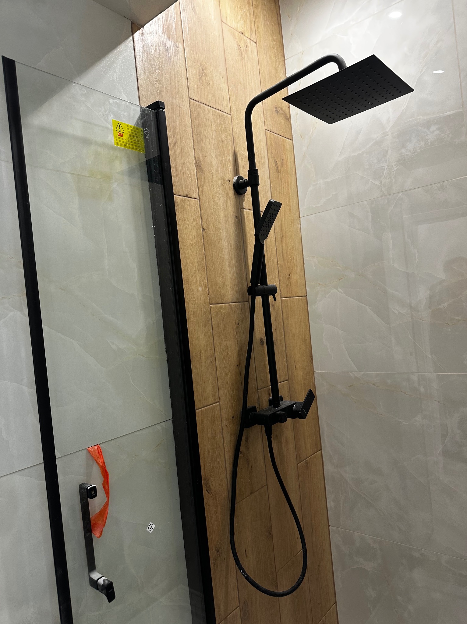 Nowoczesny zestaw natryskowy w czarnym kolorze zamontowany na ścianie wyłożonej płytkami imitującymi drewno, widoczna kabina prysznicowa z czarnymi profilami, fragment naklejki ostrzegawczej...