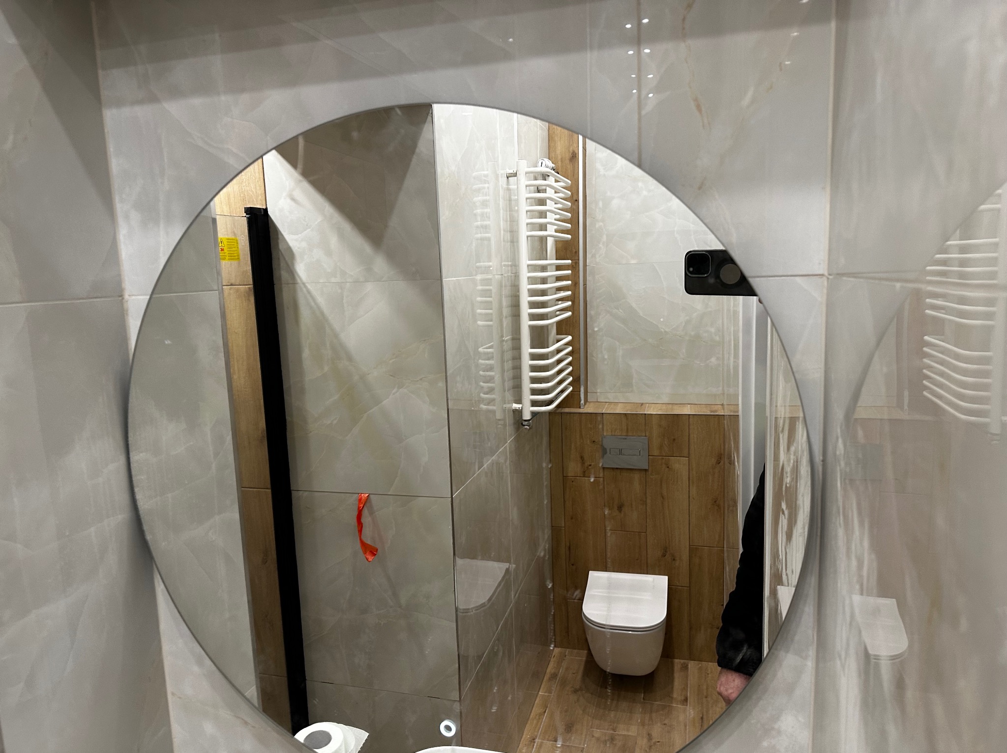 Owalne lustro z podświetleniem LED odbija fragment wykończonej łazienki z beżowymi płytkami imitującymi marmur, wiszącą toaletą, grzejnikiem drabinkowym i drewnopodobnymi panelami ściennymi.