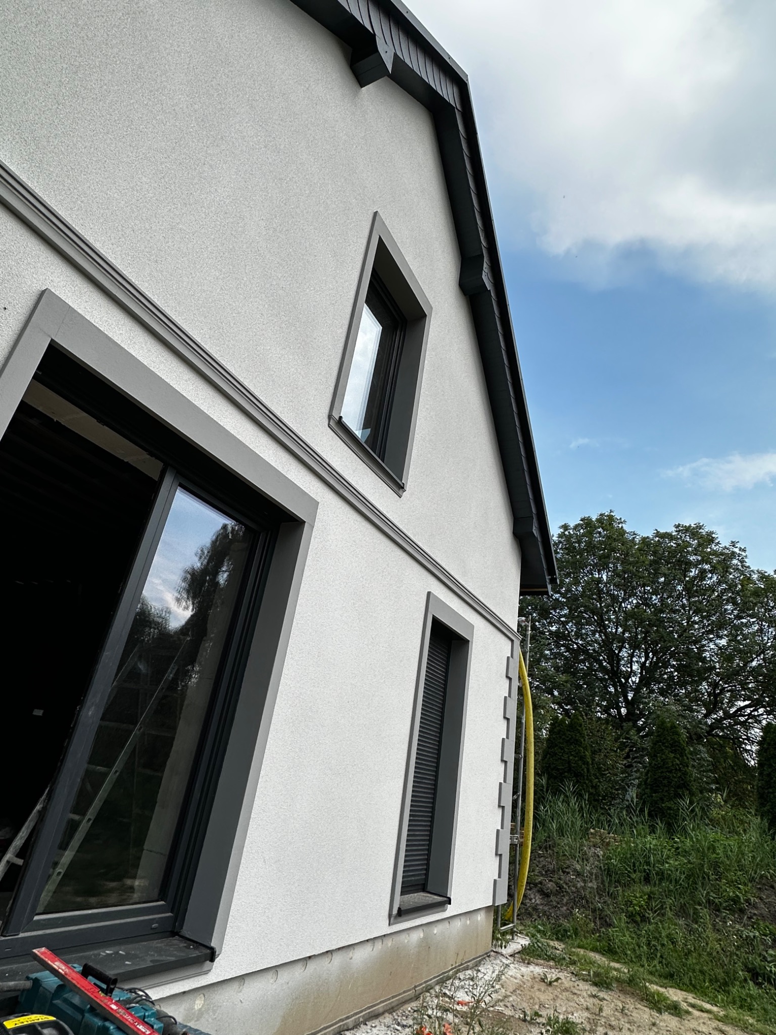 Jasnoszara elewacja domu z ciemnymi obramowaniami okien i dachu, widok z dołu ukazujący fragment drabiny oparty o ścianę i narzędzia u podstawy, błękitne niebo w tle.