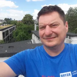 Mężczyzna na dachu z pokryciem bitumicznym, w tle budynki i zieleń. Widoczny komin i fragment dachu. Uśmiechnięta twarz, niebieska koszulka z logo firmy.