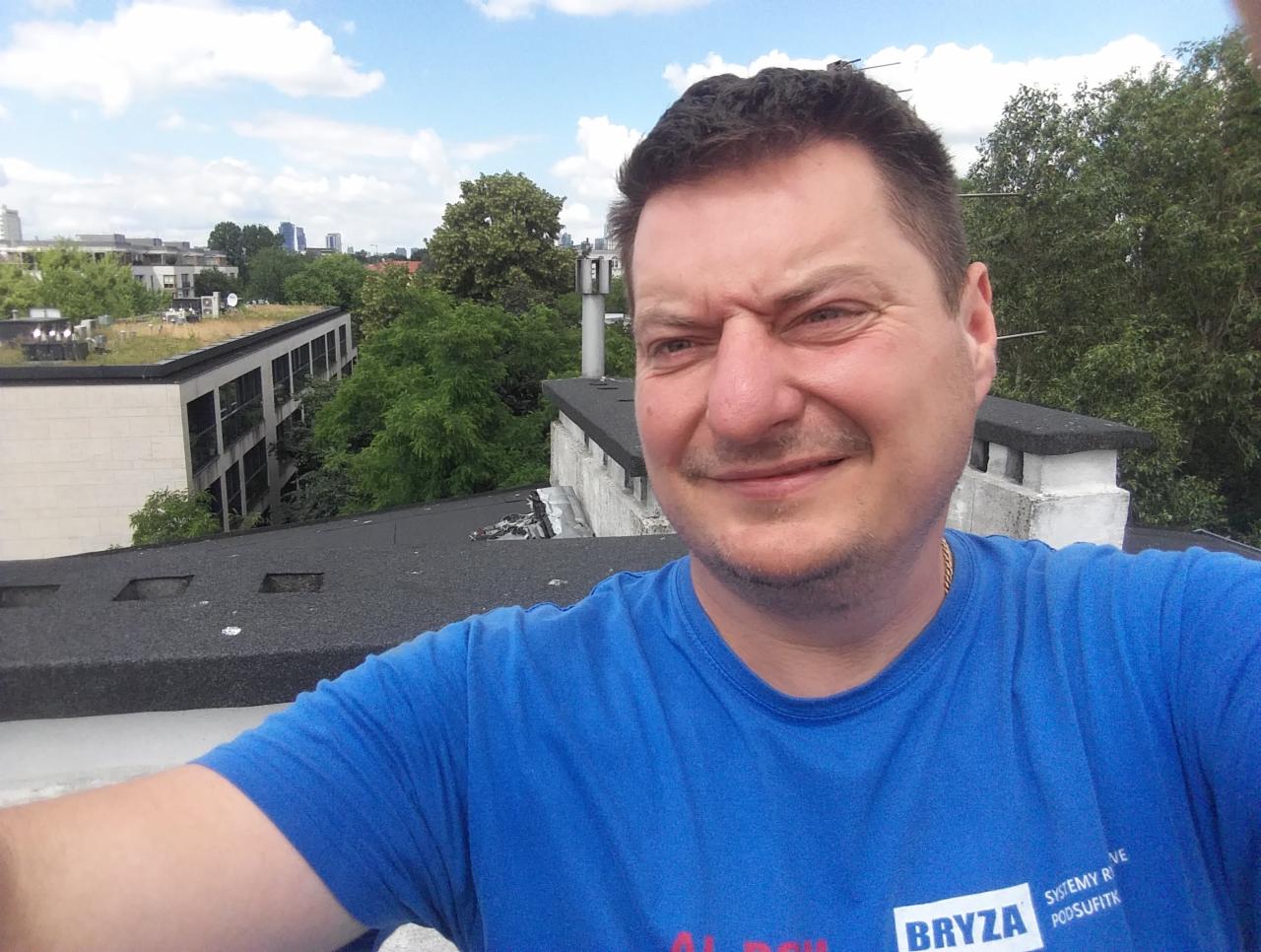 Mężczyzna na dachu z pokryciem bitumicznym, w tle budynki i zieleń. Widoczny komin i fragment dachu. Uśmiechnięta twarz, niebieska koszulka z logo firmy.