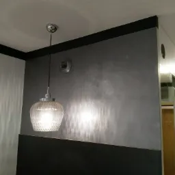 Ściana w odcieniach szarości z dekoracyjną lampą wiszącą i czarnym pasem pod sufitem, widoczny fragment wentylacji.