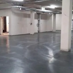 Szara, gładka posadzka przemysłowa w hali z betonowym sufitem i metalowymi rurami, oświetlona jasnym światłem, z widocznymi kolumnami nośnymi i otworami drzwiowymi w tle.