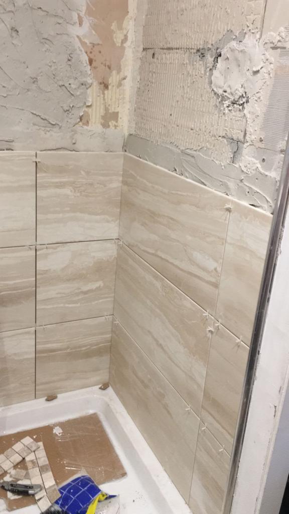 Układanie beżowych płytek imitujących drewno w rogu łazienki, widoczne krzyżyki dystansowe, fragment wanny i narzędzia na kartonie, ściana w stanie surowym.