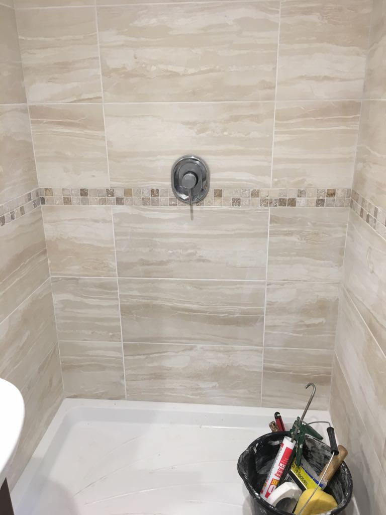 Wykończona kabina prysznicowa z beżowymi płytkami imitującymi marmur, ozdobnym pasem mozaiki i chromowaną baterią. W brodziku stoi czarne wiadro z narzędziami.