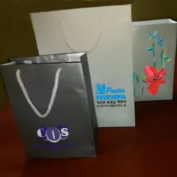 Trzy papierowe torby reklamowe z różnymi nadrukami: logo firmy COS, logo drukarni Postis i motyw kwiatowy.