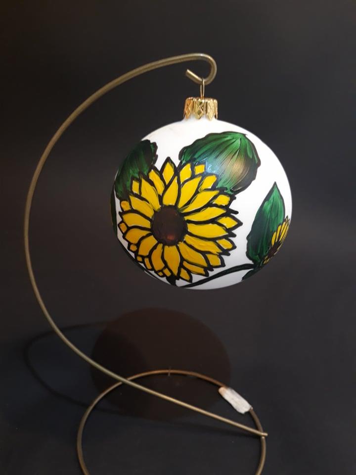 Ręcznie malowana bombka choinkowa z motywem słonecznika, zawieszona na metalowym stojaku, na ciemnym tle.