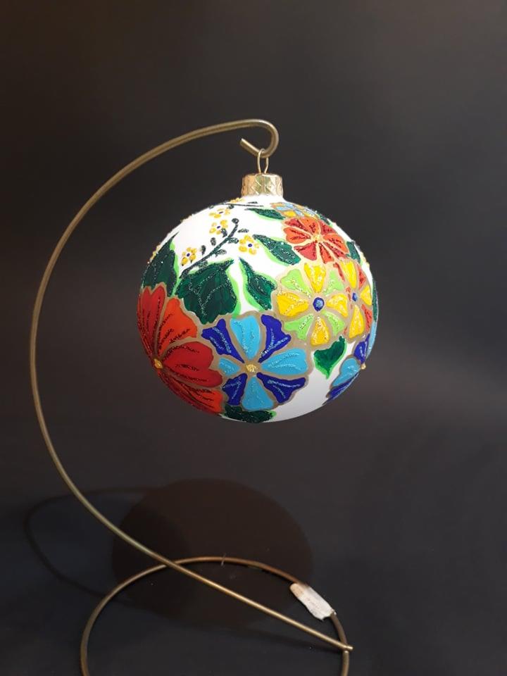 Ręcznie malowana, biała bombka świąteczna z motywem kolorowych kwiatów, zawieszona na złotym stojaku na tle czarnego tła.