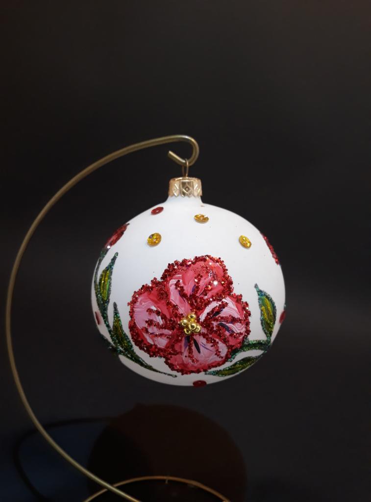 Biała bombka choinkowa ręcznie malowana z brokatowym, czerwonym kwiatem i złotymi akcentami, zawieszona na metalowym stojaku na tle czerni.