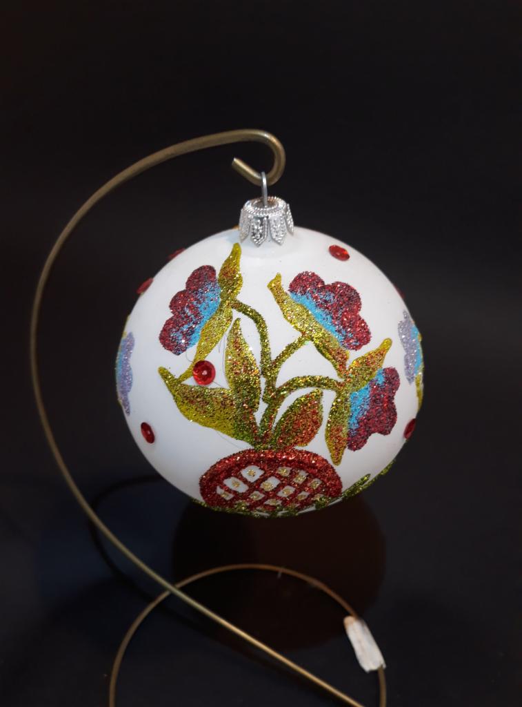 Biała bombka choinkowa z ręcznie malowanymi, brokatowymi kwiatami w odcieniach czerwieni, niebieskiego i złota, zawieszona na metalowym stojaku na tle czarnego blatu.