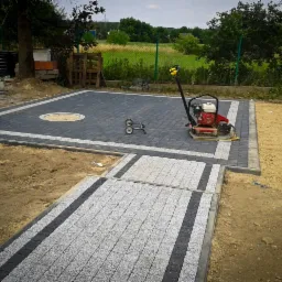 Układanie kostki brukowej: Plac z ciemnej kostki brukowej z jasną obwódką i okrągłym wzorem centralnym, fragment ścieżki z kostki granitowej w odcieniach szarości z ciemnymi pasami, zagęszczarka...