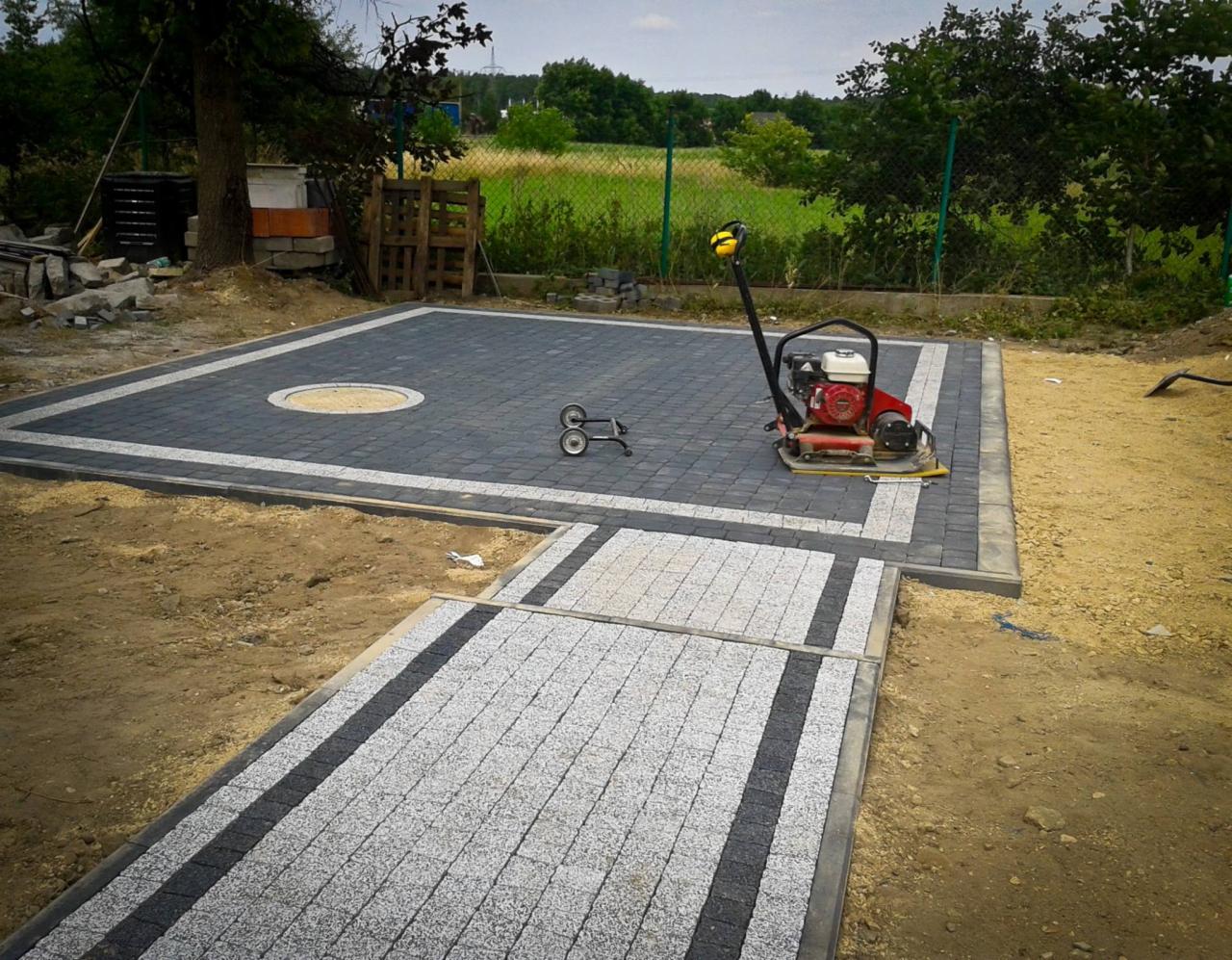 Układanie kostki brukowej: Plac z ciemnej kostki brukowej z jasną obwódką i okrągłym wzorem centralnym, fragment ścieżki z kostki granitowej w odcieniach szarości z ciemnymi pasami, zagęszczarka...