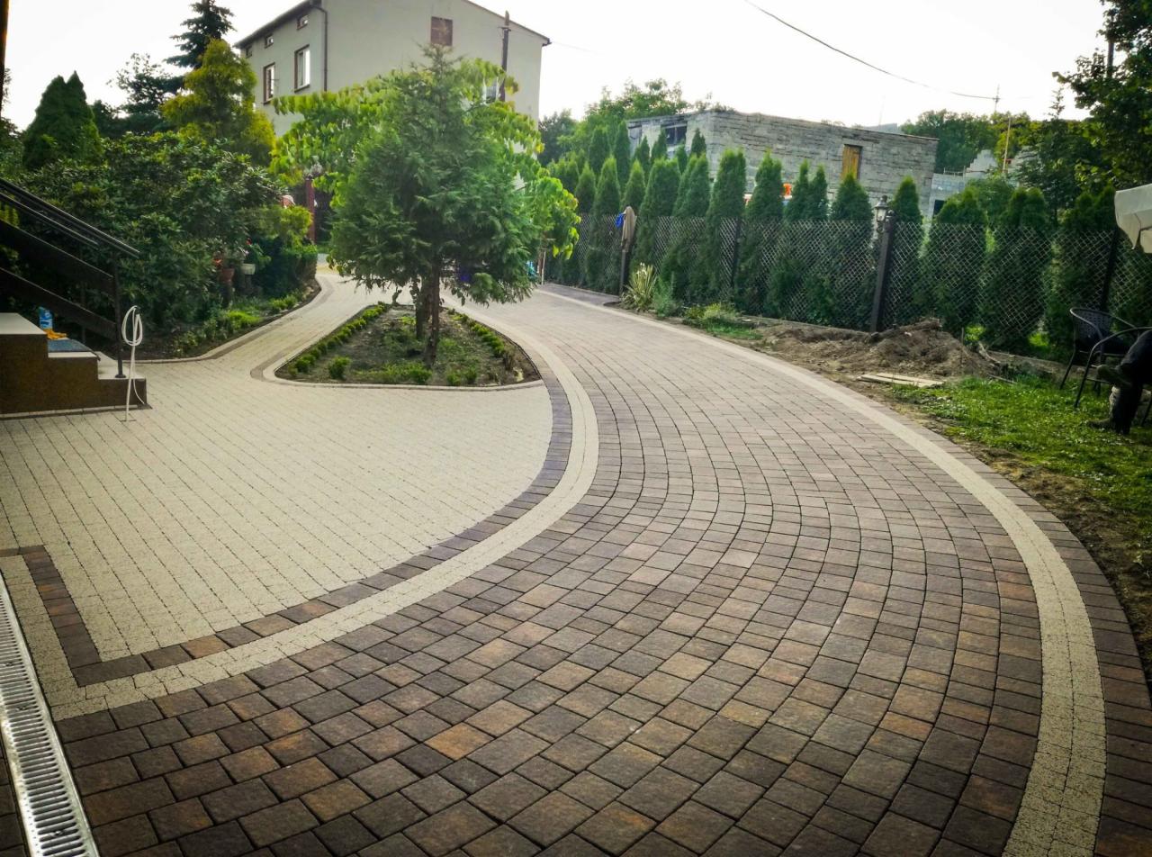 Kaskadowy układ kostki brukowej w dwóch odcieniach szarości, z ciemniejszą obwódką, tworzący ścieżkę i podjazd w ogrodzie z tujami i drzewami liściastymi w tle.