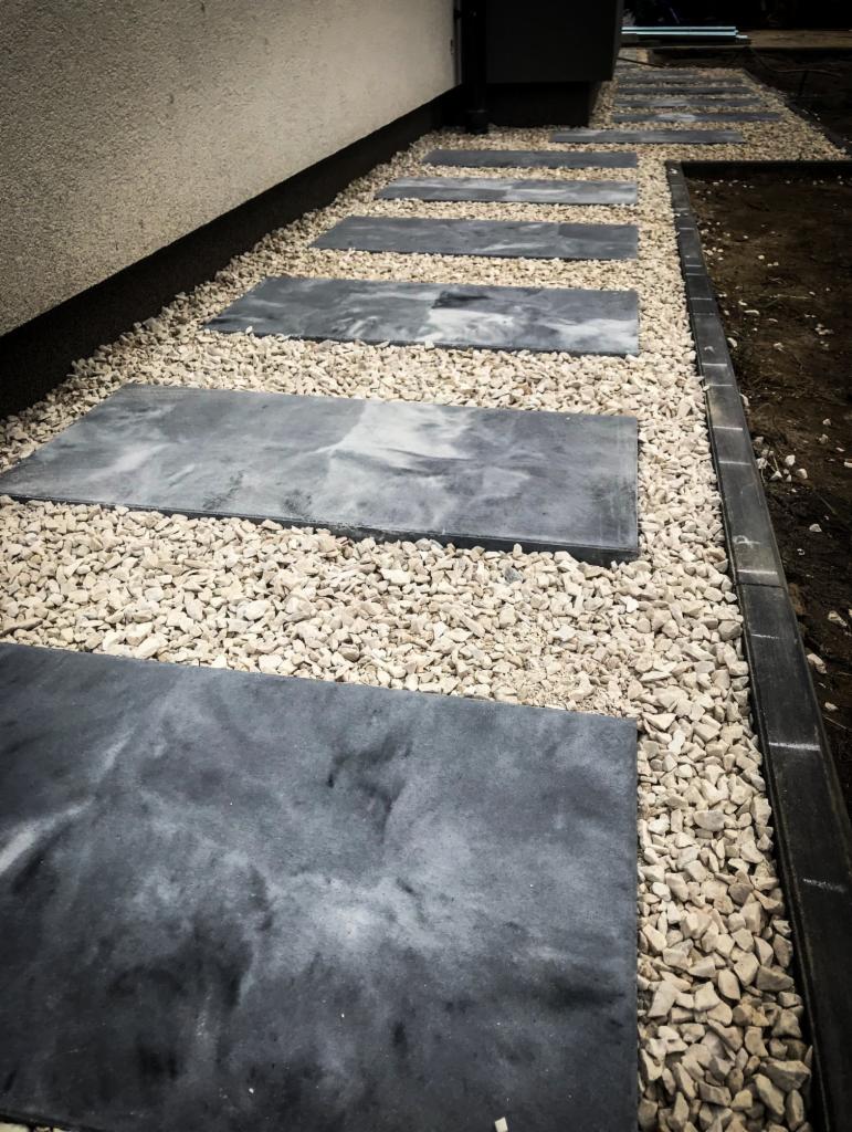 Ścieżka z prostokątnych płyt imitujących kamień, otoczona drobnym, jasnym żwirem, prowadząca wzdłuż ściany budynku.