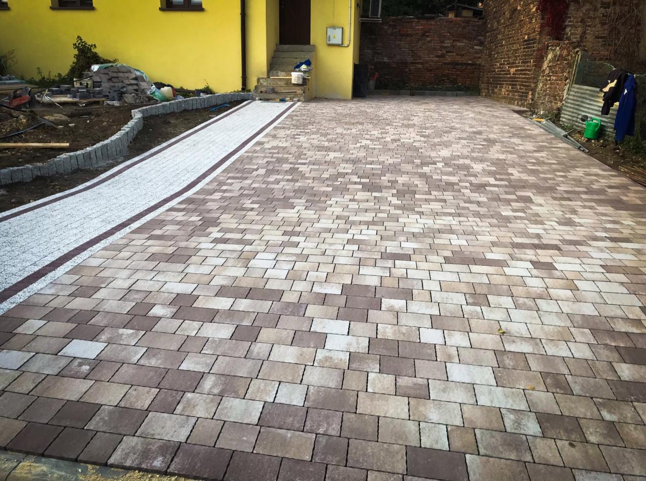 Ułożona kostka brukowa w odcieniach brązu i beżu tworząca duży plac przed żółtym budynkiem, z widocznym fragmentem chodnika z białej kostki granitowej i obrzeżami z szarego granitu.