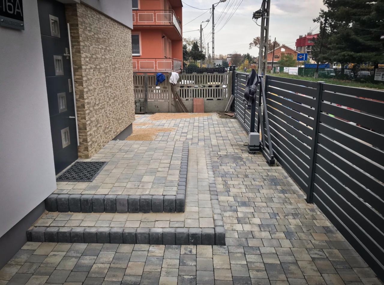 Ułożona kostka brukowa w odcieniach szarości i beżu prowadząca od schodów wejściowych do bramy posesji. Widoczne obrzeża z ciemnej kostki, szara brama panelowa i fragment elewacji budynku.