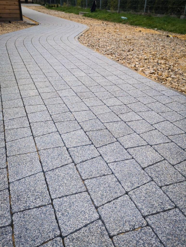 Ścieżka z szarej kostki granitowej ułożona w łuk, obok wysypany żwir, widok z góry.