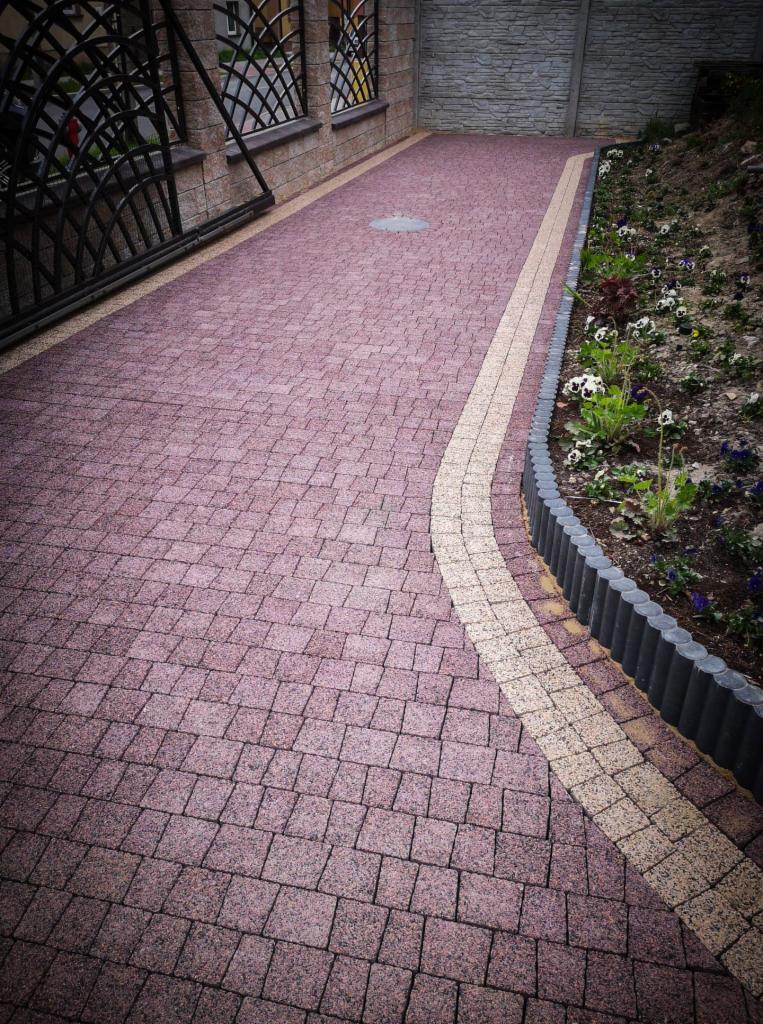 Ułożona kostka brukowa w odcieniach czerwieni z dekoracyjnym pasem z jasnej kostki, obrzeża rabat kwiatowych wzdłuż ścieżki, metalowa brama w tle.