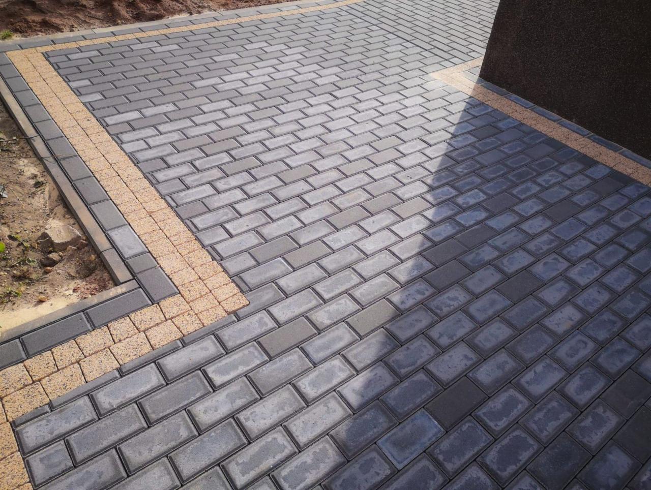 Ułożona, szara kostka brukowa z obramowaniem z kostki w kolorze piaskowym, widoczny fragment ściany budynku.