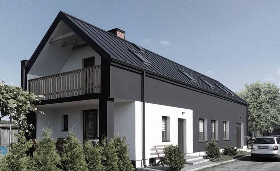 Dwukondygnacyjny dom z balkonem, wykończony w kolorach szarości i bieli, z ciemną, metalową dachówką i oknami dachowymi, otoczony zielenią.