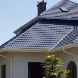 Dom z ciemnoszarą blachodachówką, widoczny komin z cegły, okno dachowe i białe rynny.