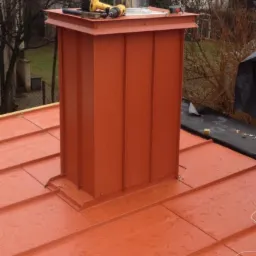 Czerwony komin z blachy na dachu, narzędzia (wiertarka, młotek) na górnej krawędzi komina, widoczny fragment pokrycia dachowego z blachy w kolorze komina.