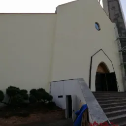 Kościół po ukończonym malowaniu elewacji na kremowy kolor, widoczne rusztowania przy wieży, schody prowadzące do wejścia głównego oraz niewielki ogródek z krzewami.