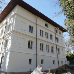 Trzypiętrowy budynek z jasną elewacją, widoczne detale architektoniczne, brązowe orynnowanie i fragment dachu, teren wokół budynku w trakcie prac wykończeniowych.