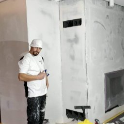 kgzabudowy@gmail.com - Mężczyzna w ubrudzonym farbą roboczym stroju i białym kapeluszu stoi na tle przygotowanej do malowania ściany z wnęką, trzymając w ręku narzędzie, z widocznym oświetleniem budowlanym...