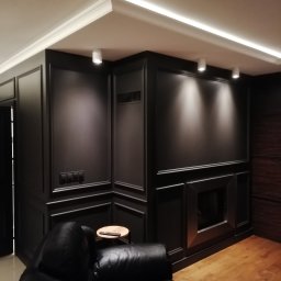 kgzabudowy@gmail.com - Eleganckie wnętrze z ciemnymi panelami ściennymi, oświetleniem punktowym i sufitem z listwami LED, widoczny fragment skórzanego fotela i drewniana podłoga.