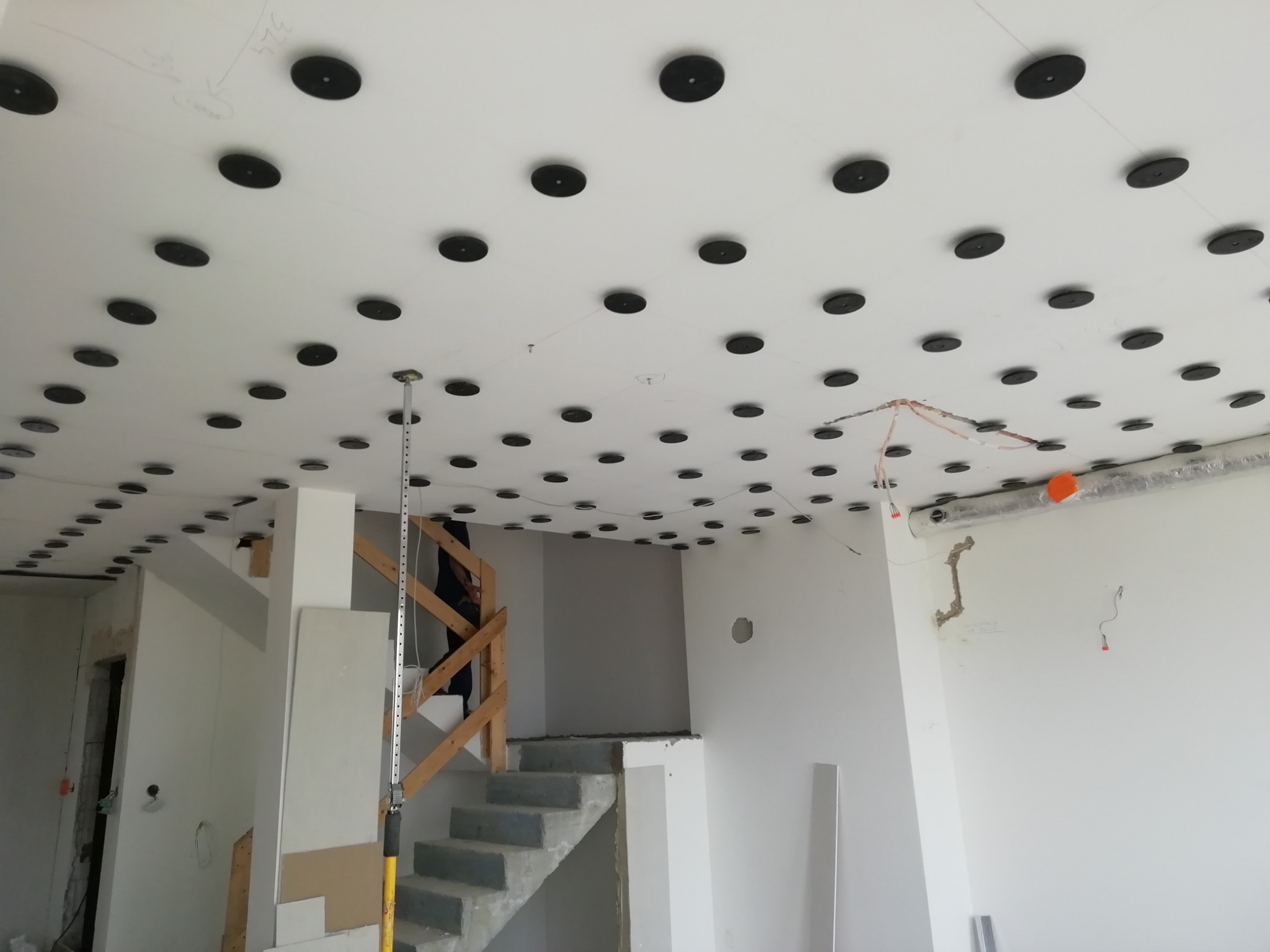 Montaż sufitu podwieszanego z widocznymi czarnymi elementami mocującymi, częściowo wykończone wnętrze z surowymi schodami i elementami instalacji elektrycznej.