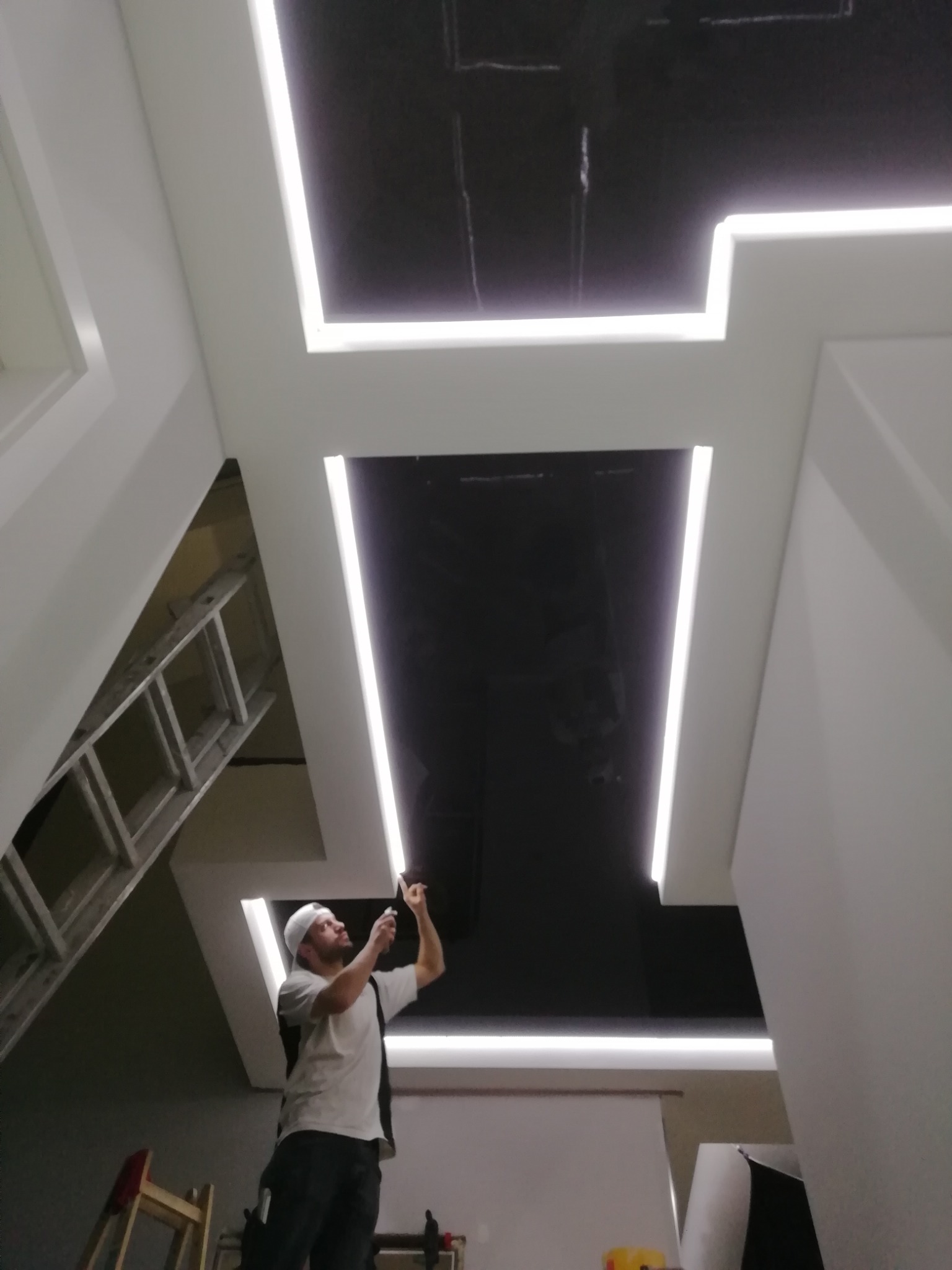 Instalacja sufitu podwieszanego z oświetleniem LED: mężczyzna na drabinie montuje listwę oświetleniową w czarnym suficie, widoczne białe panele i ukryte źródła światła.