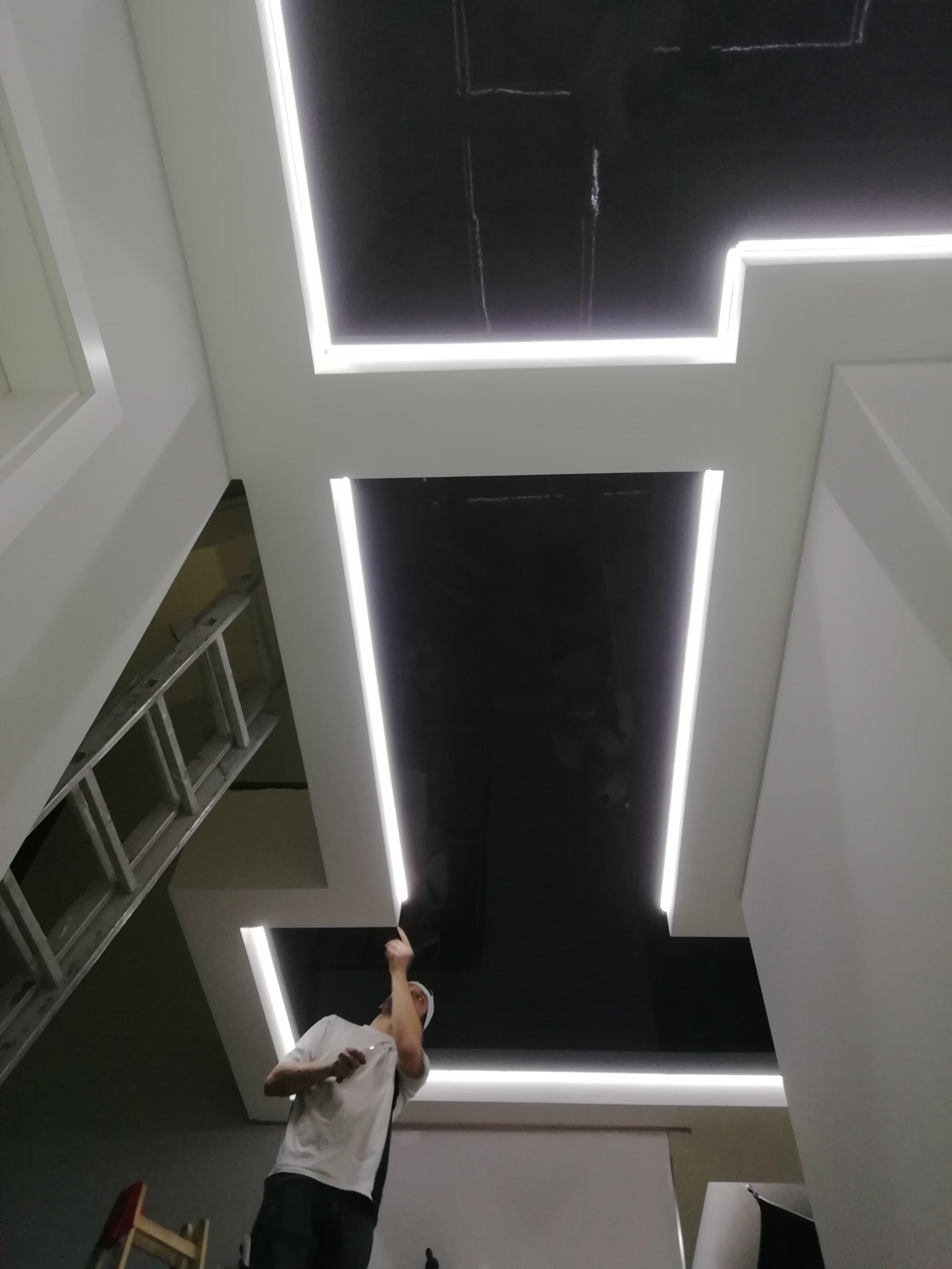 Montaż sufitu podwieszanego z czarnymi panelami i liniowym oświetleniem LED, widoczny pracownik podczas instalacji oraz drabina.