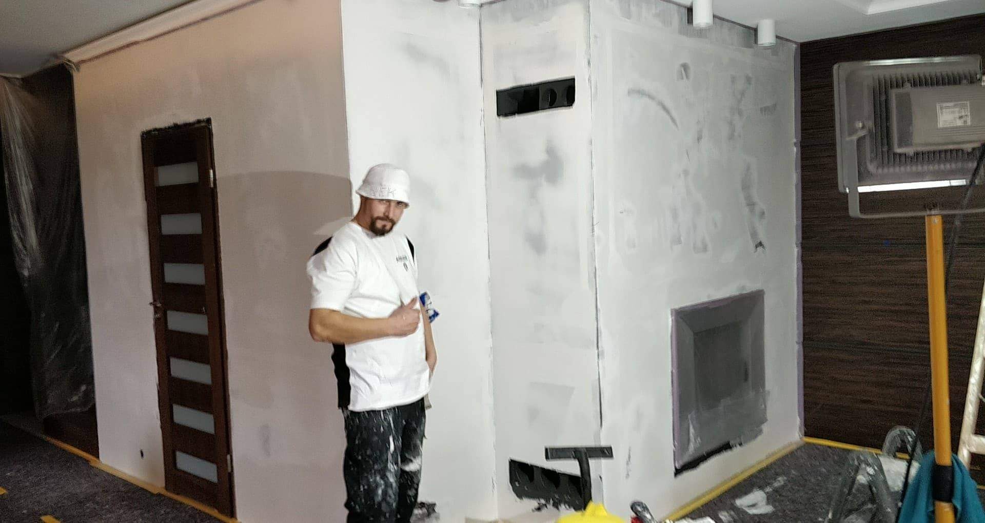 Mężczyzna w ubrudzonym farbą roboczym stroju i białym kapeluszu stoi na tle przygotowanej do malowania ściany z wnęką, trzymając w ręku narzędzie, z widocznym oświetleniem budowlanym...