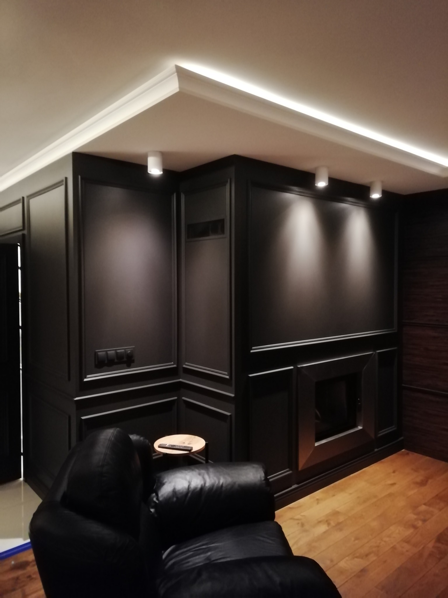 Eleganckie wnętrze z ciemnymi panelami ściennymi, oświetleniem punktowym i sufitem z listwami LED, widoczny fragment skórzanego fotela i drewniana podłoga.