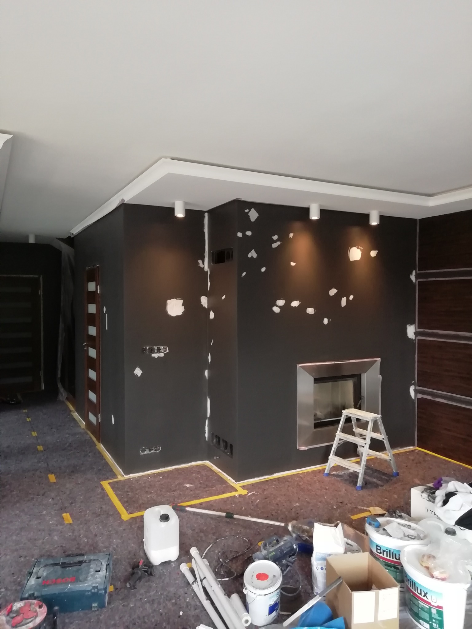 Pokój w trakcie malowania na ciemny kolor, ściany z widocznymi miejscami po szpachlowaniu, zabezpieczona podłoga, drabina przy kominku i puszki z farbą na podłodze.