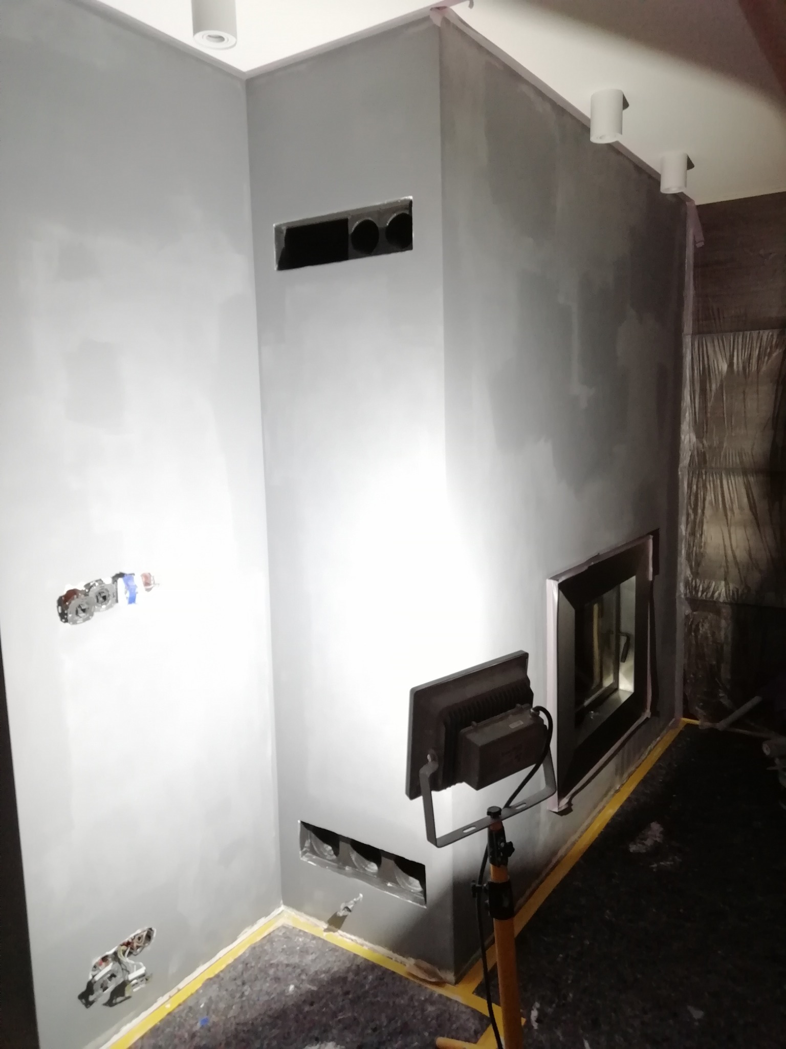 Szare ściany w trakcie malowania z przygotowanymi otworami na wentylację, widoczne gniazdka elektryczne i lampa na statywie oświetlająca obszar roboczy.