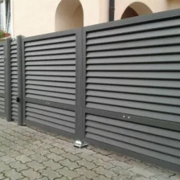 Nowoczesna, szara brama wjazdowa lamelowa z furtką, zamykana na zamek, na tle jasnej elewacji domu z kwiatami w oknie.