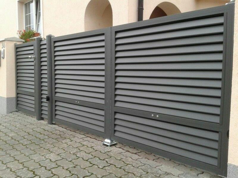 Nowoczesna, szara brama wjazdowa lamelowa z furtką, zamykana na zamek, na tle jasnej elewacji domu z kwiatami w oknie.