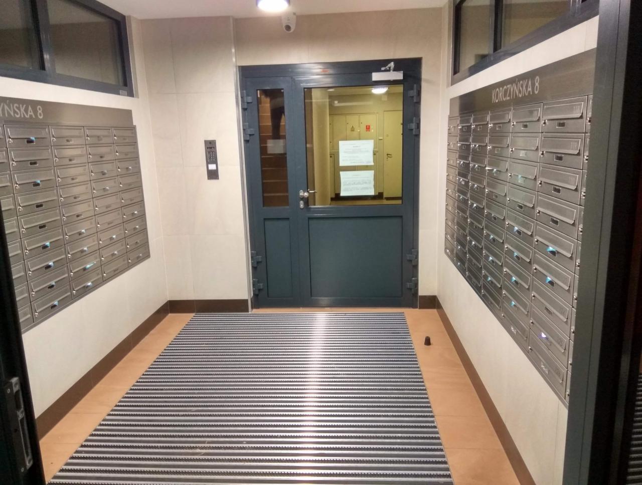 Wnętrze klatki schodowej z metalowymi skrzynkami pocztowymi po obu stronach, widoczne drzwi wejściowe z przeszklonym panelem i wycieraczka w paski.