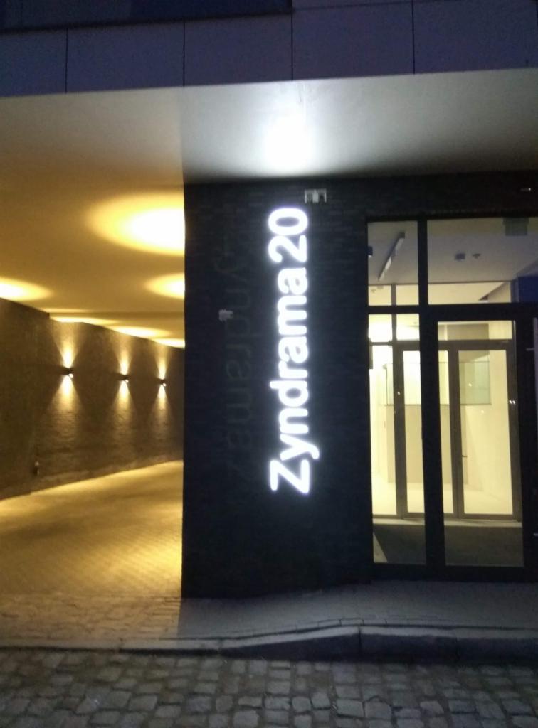 Oświetlony wjazd do garażu z podświetlanym numerem budynku Zyndrama 20, widoczne lampy ścienne i szklane drzwi wejściowe.