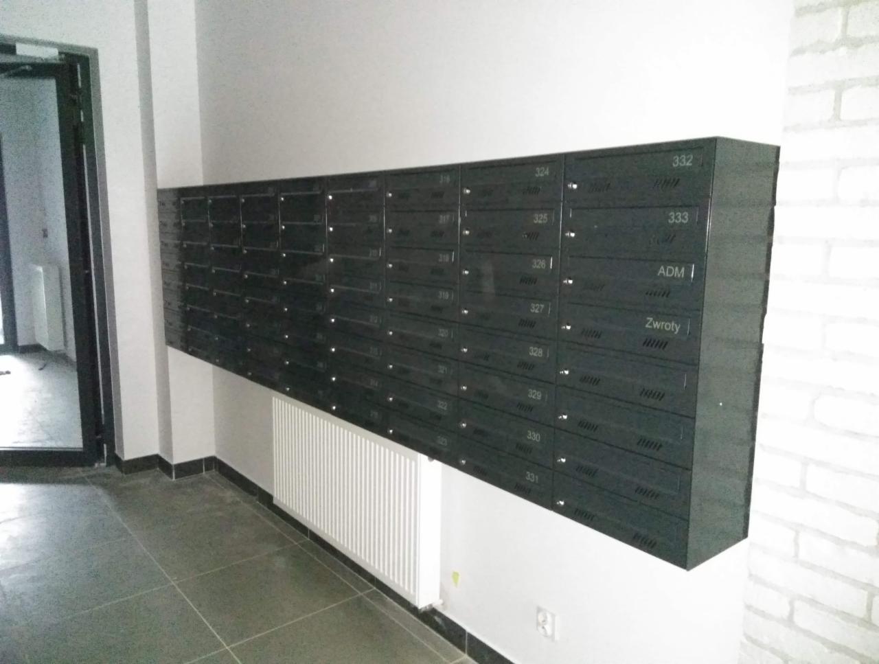 Rząd czarnych skrzynek pocztowych wewnątrz budynku, numery od 324 do 333, napisy 'ADM' i 'Zwroty' na niektórych skrzynkach, biały grzejnik pod spodem, szara podłoga z płytek.