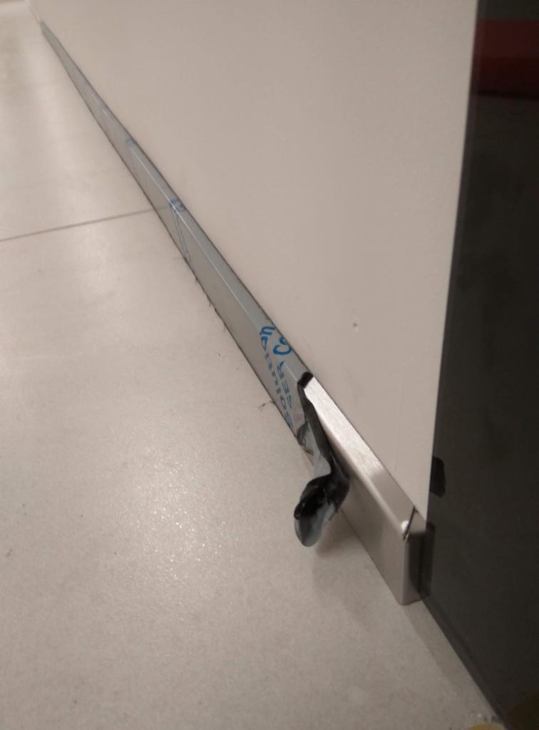 Detal montażu dolnej prowadnicy przesuwnej konstrukcji, zabezpieczonej folią ochronną, przy białej ścianie i szarej podłodze.