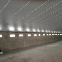 Wnętrze w trakcie budowy z rzędem małych okien w betonowej ścianie i panelowym sufitem z widocznymi mocowaniami, materiały budowlane na podłodze.