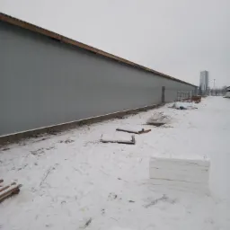 Długi, szary budynek przemysłowy z widocznym zadaszeniem, otoczony śniegiem i elementami budowlanymi.