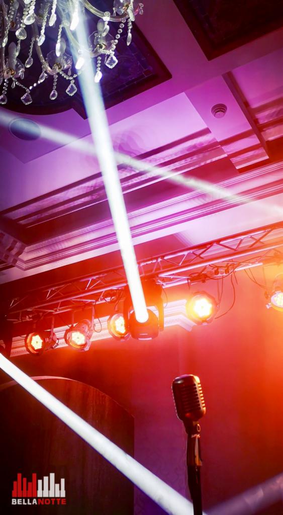Scena oświetlona reflektorami z mikrofonem w stylu retro na pierwszym planie, widoczny żyrandol i logo 'Bella Notte' w lewym dolnym rogu.