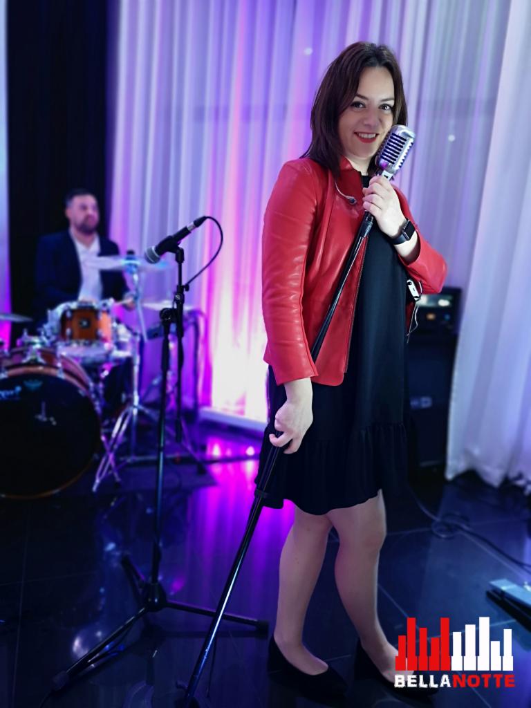 Wokalistka w czerwonej skórzanej kurtce trzyma mikrofon na scenie, w tle perkusista i fioletowe oświetlenie.