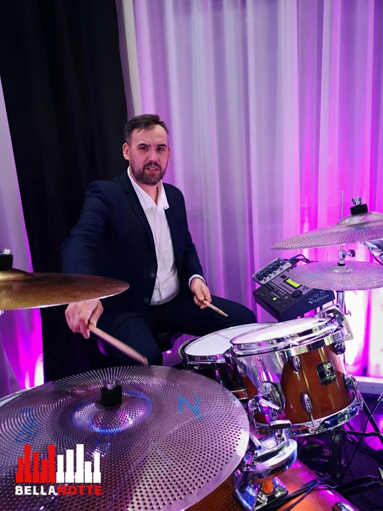 Perkusista w garniturze grający na perkusji na scenie z fioletowym oświetleniem i logo zespołu BellaNotte na talerzu perkusyjnym.