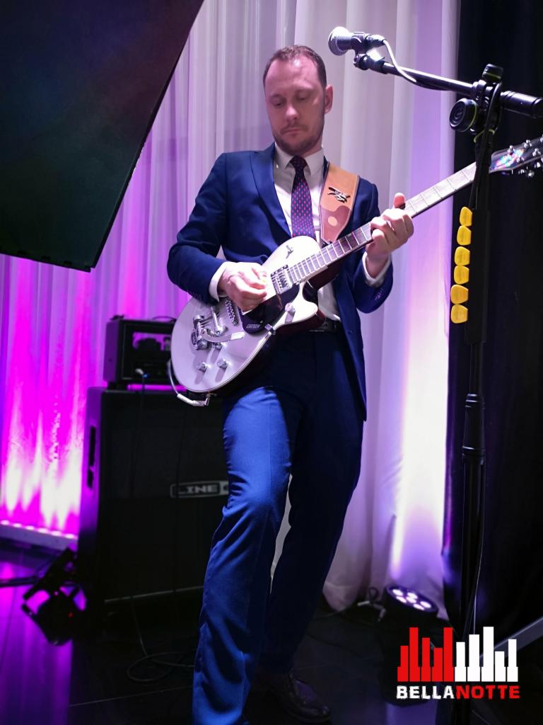 Gitarzysta w niebieskim garniturze gra na białej gitarze elektrycznej podczas występu zespołu BellaNotte, z widocznym wzmacniaczem i mikrofonem.